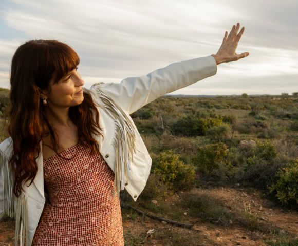 A woman with long brown hair wears a fringed white jacket and a red and white checkered dress as she holds her hand up to block the sun in a dry, brush-covered landscape under a cloudy sky.