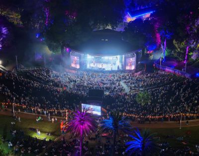Aerial view of an outdoor amphitheater at night, with a band performing on a brightly lit stage and a large audience seated on a grassy hill. Colorful lights illuminate the trees and palm trees surrounding the venue.