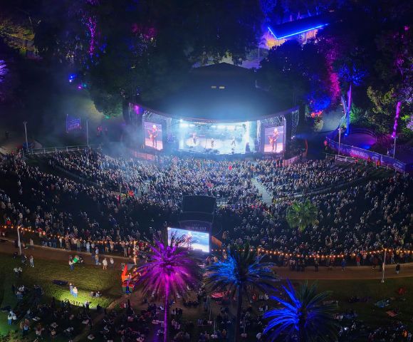 Aerial view of an outdoor amphitheater at night, with a band performing on a brightly lit stage and a large audience seated on a grassy hill. Colorful lights illuminate the trees and palm trees surrounding the venue.