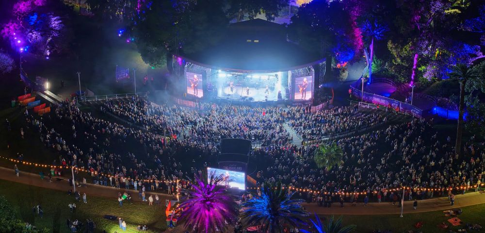 Aerial view of an outdoor amphitheater at night, with a band performing on a brightly lit stage and a large audience seated on a grassy hill. Colorful lights illuminate the trees and palm trees surrounding the venue.
