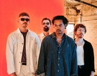 Four men stand in a line against an orange backdrop, with one man in the foreground wearing a denim shirt and another in a white jacket.