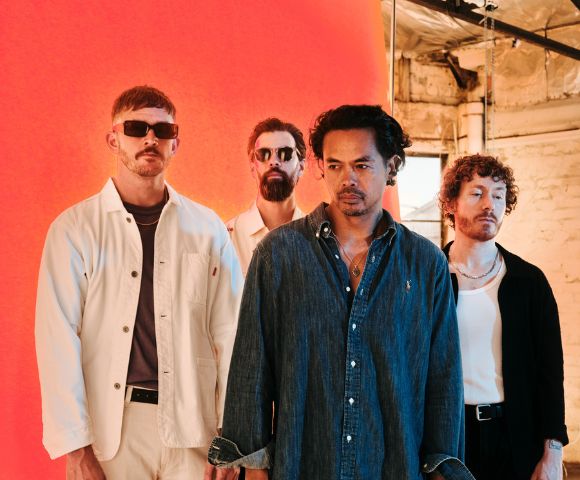 Four men stand in a line against an orange backdrop, with one man in the foreground wearing a denim shirt and another in a white jacket.