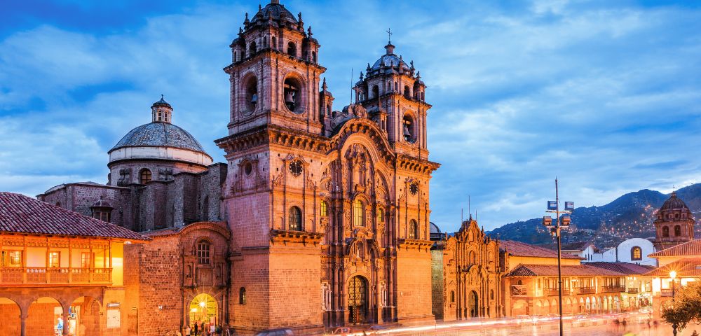 Historic cathedral in Cusco, Peru, illuminated at dusk. The baroque architecture features ornate towers against a blue sky, evoking a serene atmosphere.