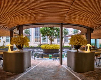 Modern outdoor seating area with a curved wooden ceiling, concrete counters with lamps and floral arrangements, and a courtyard with trees and seating.