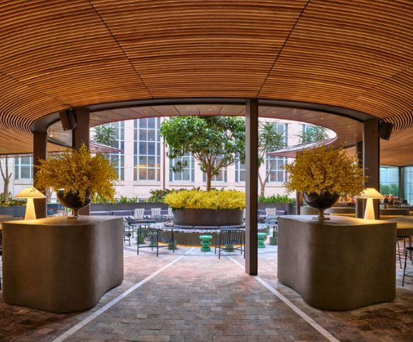 Modern outdoor seating area with a curved wooden ceiling, concrete counters with lamps and floral arrangements, and a courtyard with trees and seating.