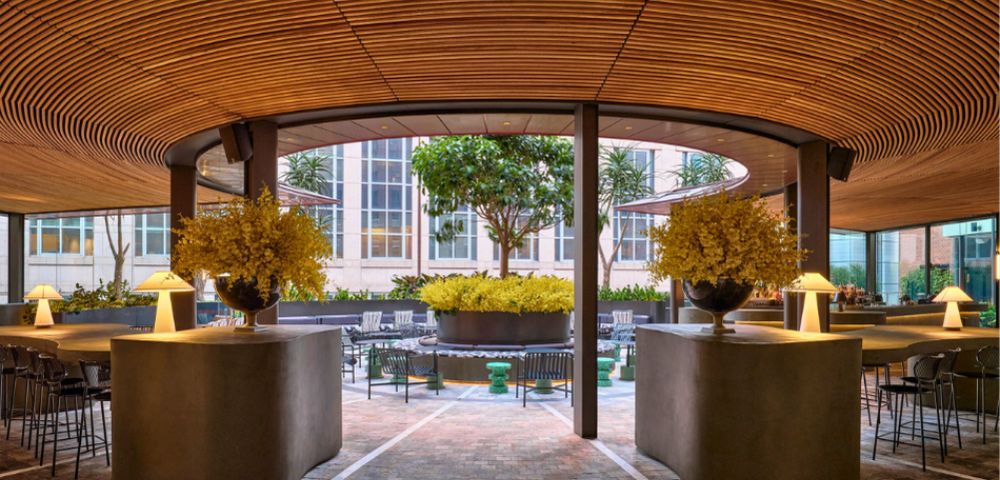 Modern outdoor seating area with a curved wooden ceiling, concrete counters with lamps and floral arrangements, and a courtyard with trees and seating.