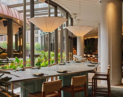 A long, stone-topped table with place settings for dining in a modern restaurant with large windows looking out onto lush greenery.