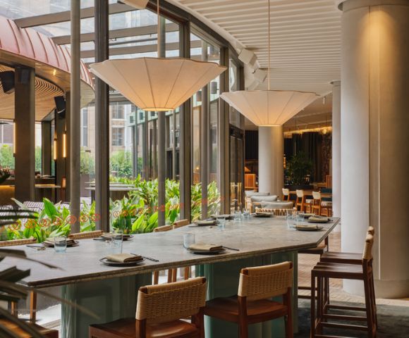 A long, stone-topped table with place settings for dining in a modern restaurant with large windows looking out onto lush greenery.