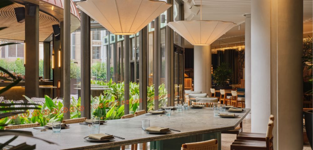 A long, stone-topped table with place settings for dining in a modern restaurant with large windows looking out onto lush greenery.