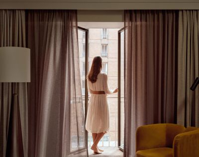 Woman in a white dress with a brown belt opens tall glass doors to reveal a brick building with balconies.