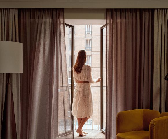 Woman in a white dress with a brown belt opens tall glass doors to reveal a brick building with balconies.