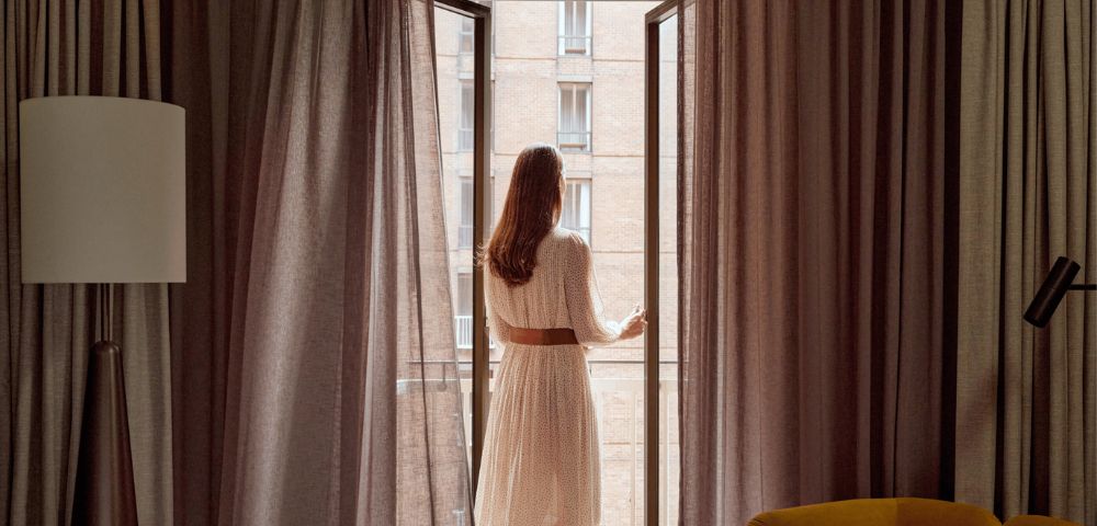 Woman in a white dress with a brown belt opens tall glass doors to reveal a brick building with balconies.