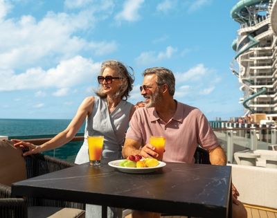 Elderly couple enjoying drinks on a sunny deck with ocean view, fruit plate, and modern building in background, evoking relaxation and joy.