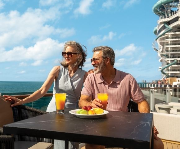 Elderly couple enjoying drinks on a sunny deck with ocean view, fruit plate, and modern building in background, evoking relaxation and joy.