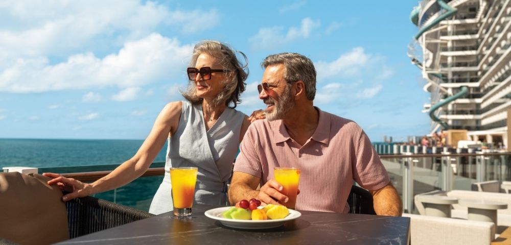 Elderly couple enjoying drinks on a sunny deck with ocean view, fruit plate, and modern building in background, evoking relaxation and joy.