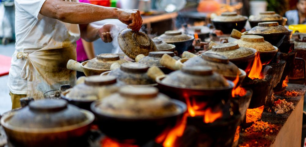 A chef stirring clay pots over an open flame in a bustling street food market, creating delicious traditional dishes.