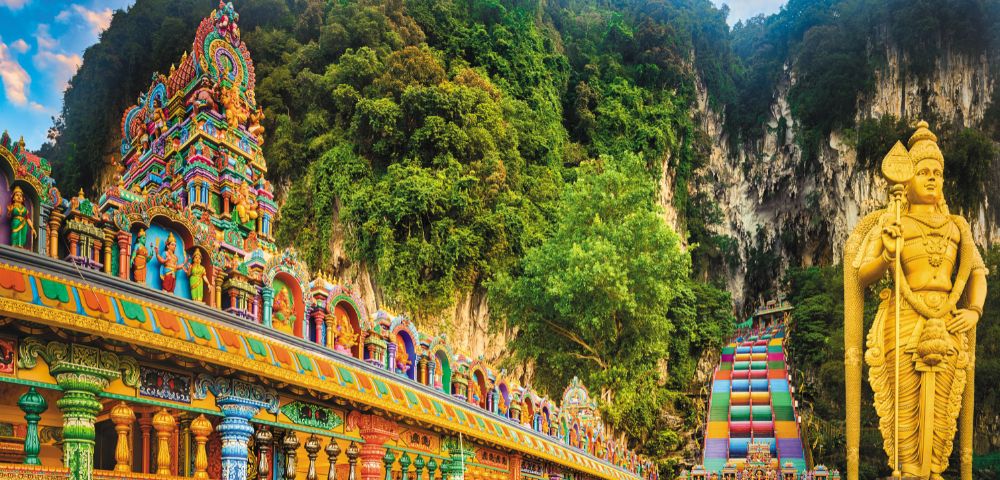 Vibrant Batu Caves temple complex with colorful architecture surrounded by lush greenery and towering limestone cliffs in Malaysia.