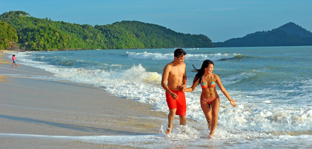 A couple holding hands and walking along the beach shoreline with waves and green forested hills in the background.