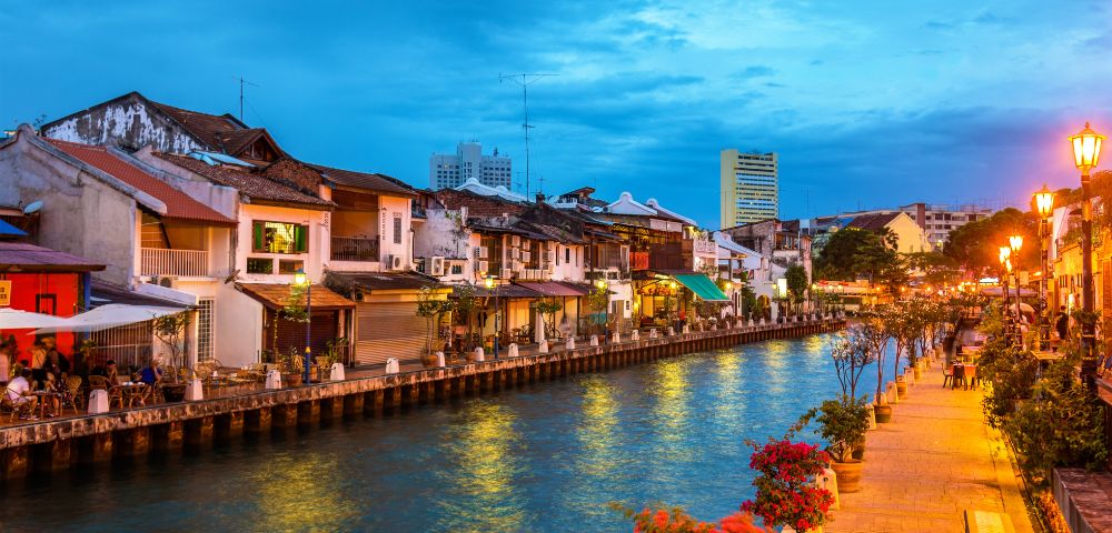 Evening view of a river lined with old buildings and street lamps, with outdoor seating and vibrant flowers along the walkway.