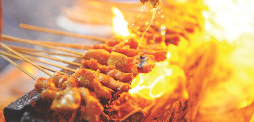 Skewers of seasoned meat grilling over an open flame with oil or marinade being drizzled on top.