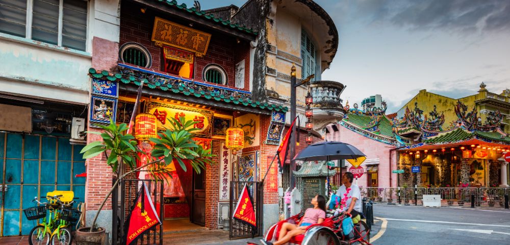A traditional rickshaw carrying two people travels down a street lined with ornate, colorful buildings adorned with lanterns.