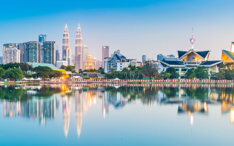 Reflective cityscape of Kuala Lumpur at twilight, featuring the Petronas Twin Towers, a television tower, and modern skyscrapers reflected in a calm lake.