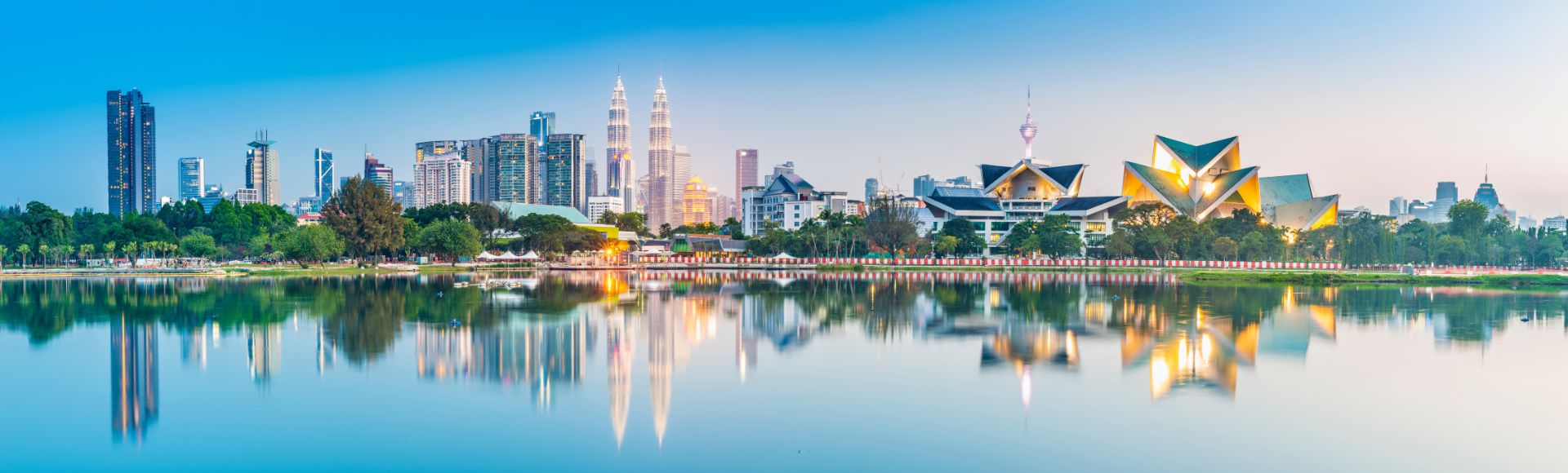 Reflective cityscape of Kuala Lumpur at twilight, featuring the Petronas Twin Towers, a television tower, and modern skyscrapers reflected in a calm lake.