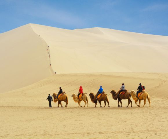 A caravan of camels with riders traverses a vast, rolling sand dune under a clear blue sky, with small figures ascending a distant dune.