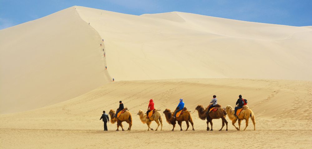 A caravan of camels with riders traverses a vast, rolling sand dune under a clear blue sky, with small figures ascending a distant dune.