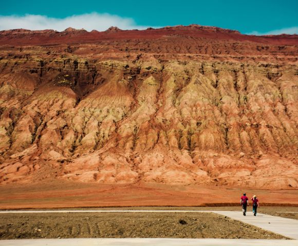 Two people walk on a paved path in front of a vast, multi-layered red and tan rock formation under a bright blue sky.