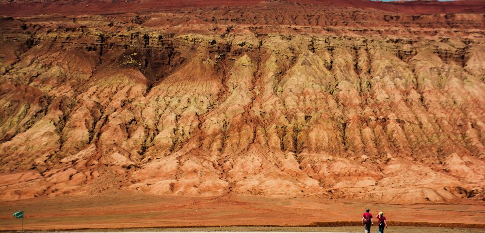 Two people walk on a paved path in front of a vast, multi-layered red and tan rock formation under a bright blue sky.