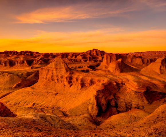 A wide, arid landscape of eroded rock formations bathed in the warm, orange glow of sunset.