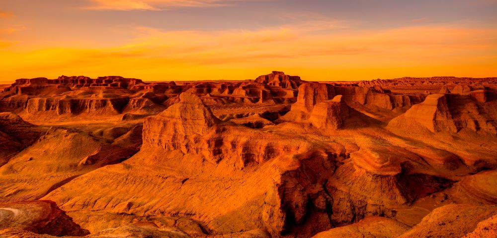 A wide, arid landscape of eroded rock formations bathed in the warm, orange glow of sunset.