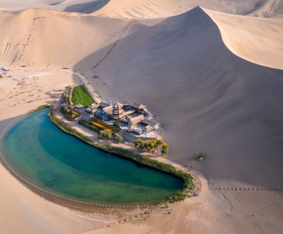 Aerial view of the Crescent Lake and the Singing Sand Dunes with traditional Chinese architecture, Lush green vegetation surrounds the oasis in a vast desert landscape.