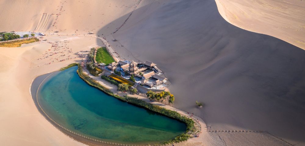 Aerial view of the Crescent Lake and the Singing Sand Dunes with traditional Chinese architecture, Lush green vegetation surrounds the oasis in a vast desert landscape.
