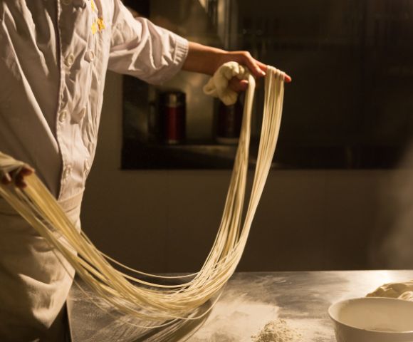 Chef stretching dough into long, thin noodles over a stainless steel counter with steam rising in the background.