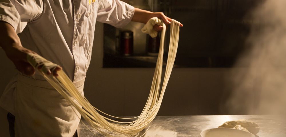 Chef stretching dough into long, thin noodles over a stainless steel counter with steam rising in the background.