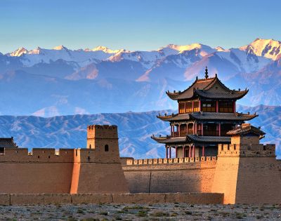 A traditional Chinese fortress wall with watchtowers and a multi-tiered pavilion under a clear blue sky, with snow-capped mountains in the background.
