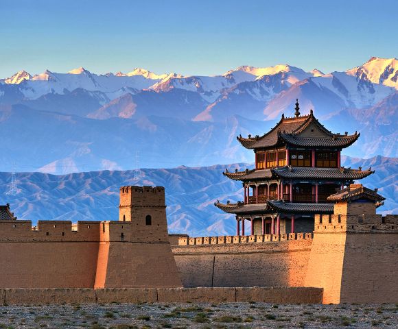 A traditional Chinese fortress wall with watchtowers and a multi-tiered pavilion under a clear blue sky, with snow-capped mountains in the background.