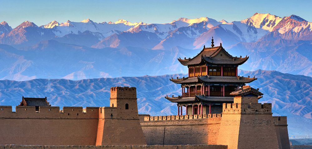A traditional Chinese fortress wall with watchtowers and a multi-tiered pavilion under a clear blue sky, with snow-capped mountains in the background.