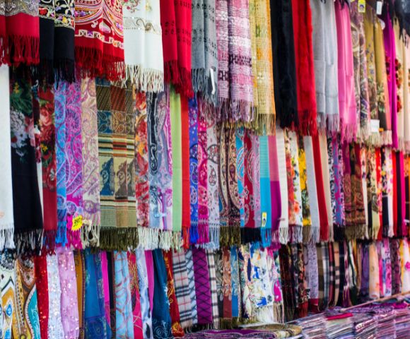 A vibrant display of intricately patterned scarves hangs in a market stall, featuring diverse designs like paisley, floral, and geometric prints in a multitude of colors.