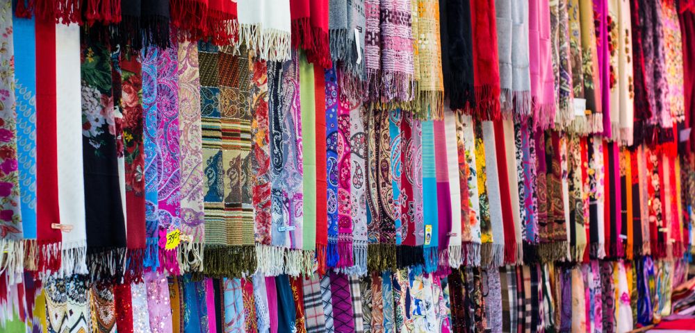 A vibrant display of intricately patterned scarves hangs in a market stall, featuring diverse designs like paisley, floral, and geometric prints in a multitude of colors.