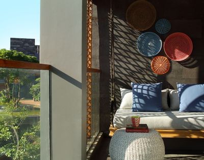 Sunlit cozy seating area with blue cushions, woven wall decor, and a white knitted ottoman next to a glass balcony overlooking greenery.