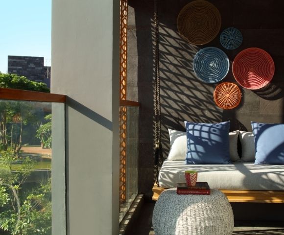 Sunlit cozy seating area with blue cushions, woven wall decor, and a white knitted ottoman next to a glass balcony overlooking greenery.
