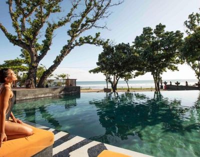Woman in swimsuit sitting by an infinity pool with trees, ocean, and clear sky in the background.
