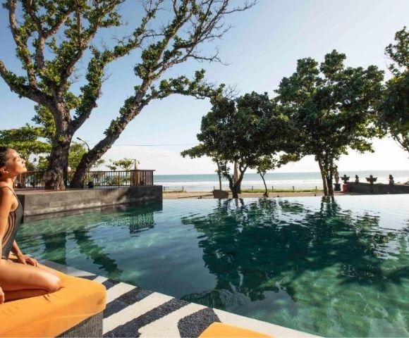 Woman in swimsuit sitting by an infinity pool with trees, ocean, and clear sky in the background.
