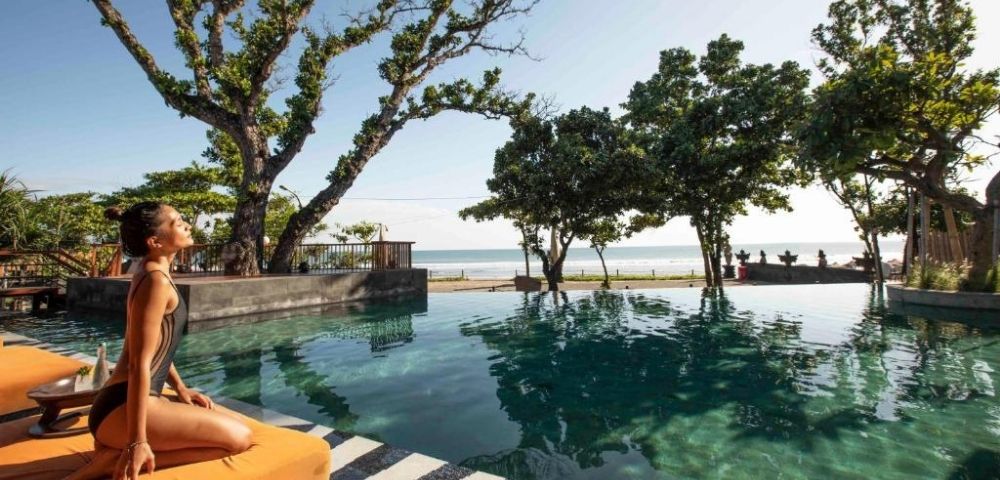 Woman in swimsuit sitting by an infinity pool with trees, ocean, and clear sky in the background.