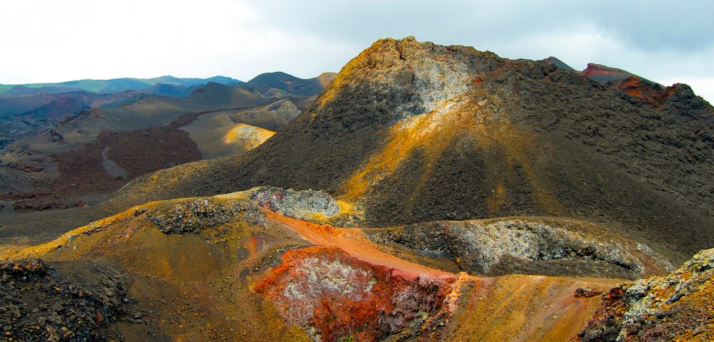 Volcanic landscape with multicolored rocky terrain and hills under a cloudy sky.