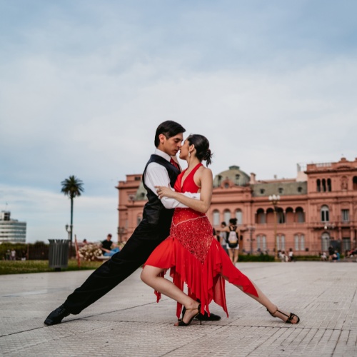 A couple performs a passionate tango on a plaza, with a historic pink building in the background. The woman wears a red dress, while the man is in black.