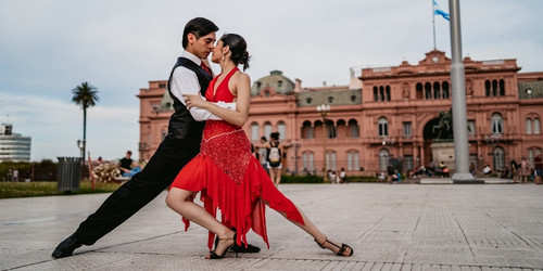 A couple performs a passionate tango on a plaza, with a historic pink building in the background. The woman wears a red dress, while the man is in black.